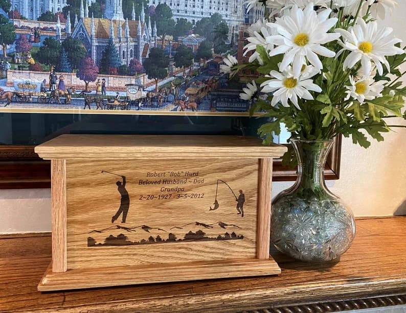 Handcrafted white oak cremation urn with custom laser engravings of a golfer, a fisherman, mountains, and trees for a sports tribute
